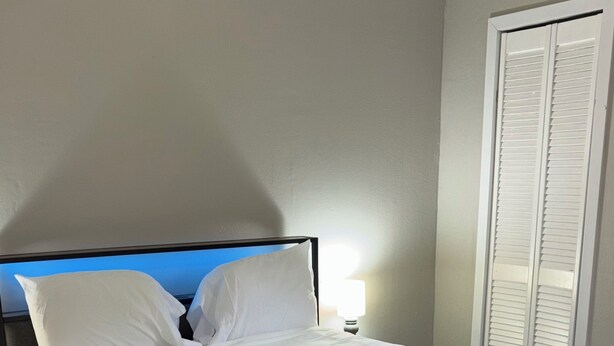 Una cama bien tendida con sábanas blancas y un edredón negro, una lámpara de noche y suelo de madera.