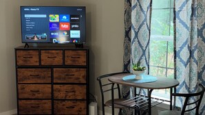Una habitación con un armario de madera, un televisor mostrando la interfaz de un servicio de streaming, una mesa de comedor con sillas y una ventana con cortinas estampadas.