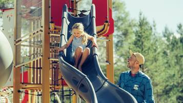 A child sliding down a playground slide with a man standing beside her.