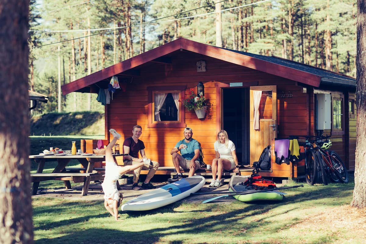 A wooden cabin with a porch, a picnic table, paddleboards, and a bicycle.