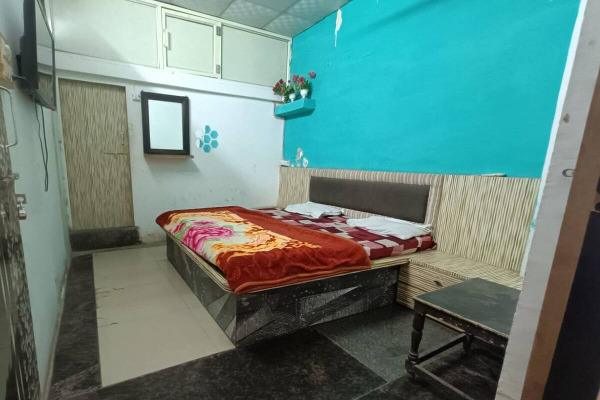 A compact bedroom with a bed, a small table, a mirror, and a wall-mounted shelf with flowers.