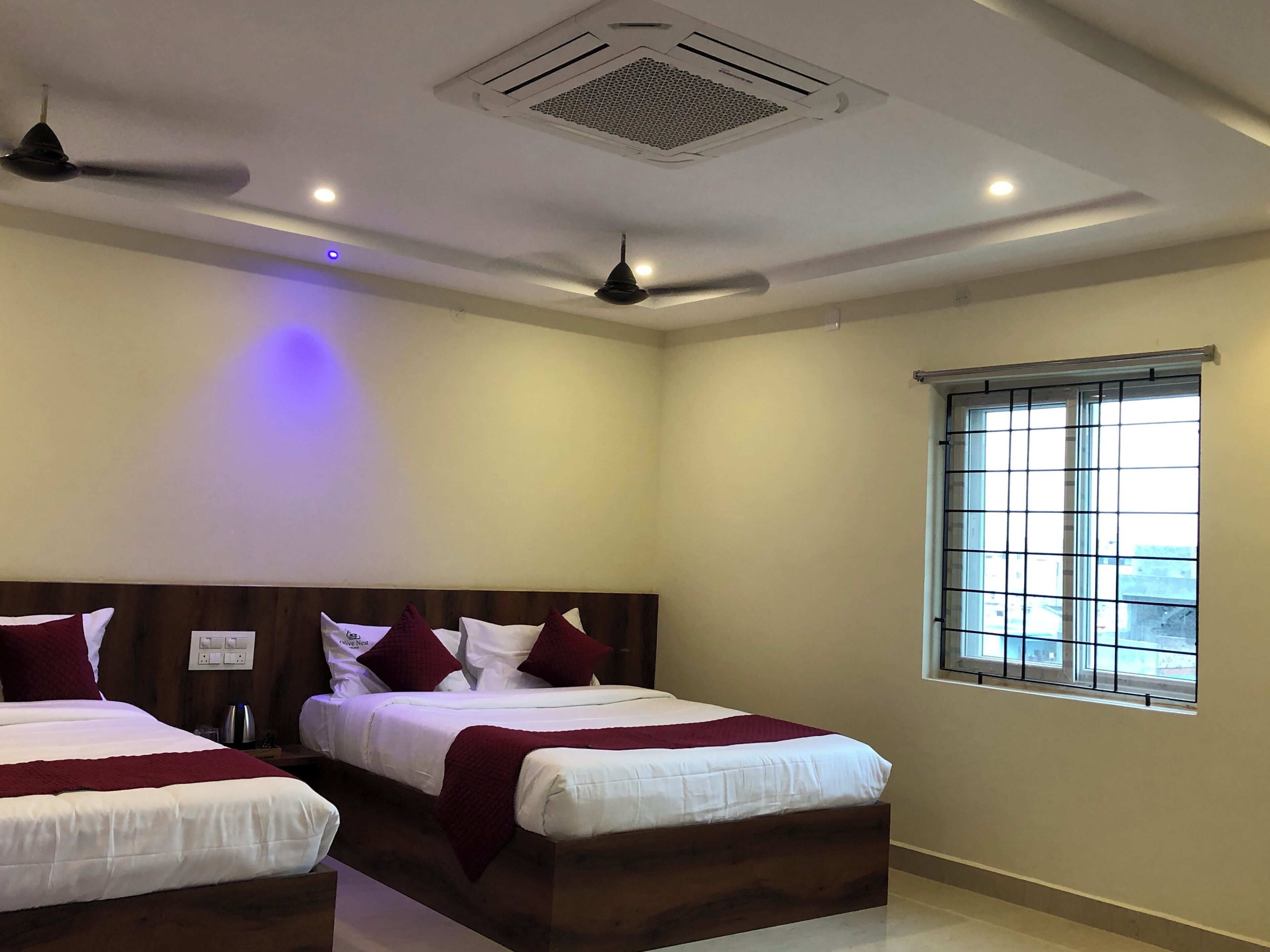 Quarto de hotel com duas camas, ventoinha no teto, ar condicionado, janela com grades e vista para o exterior.