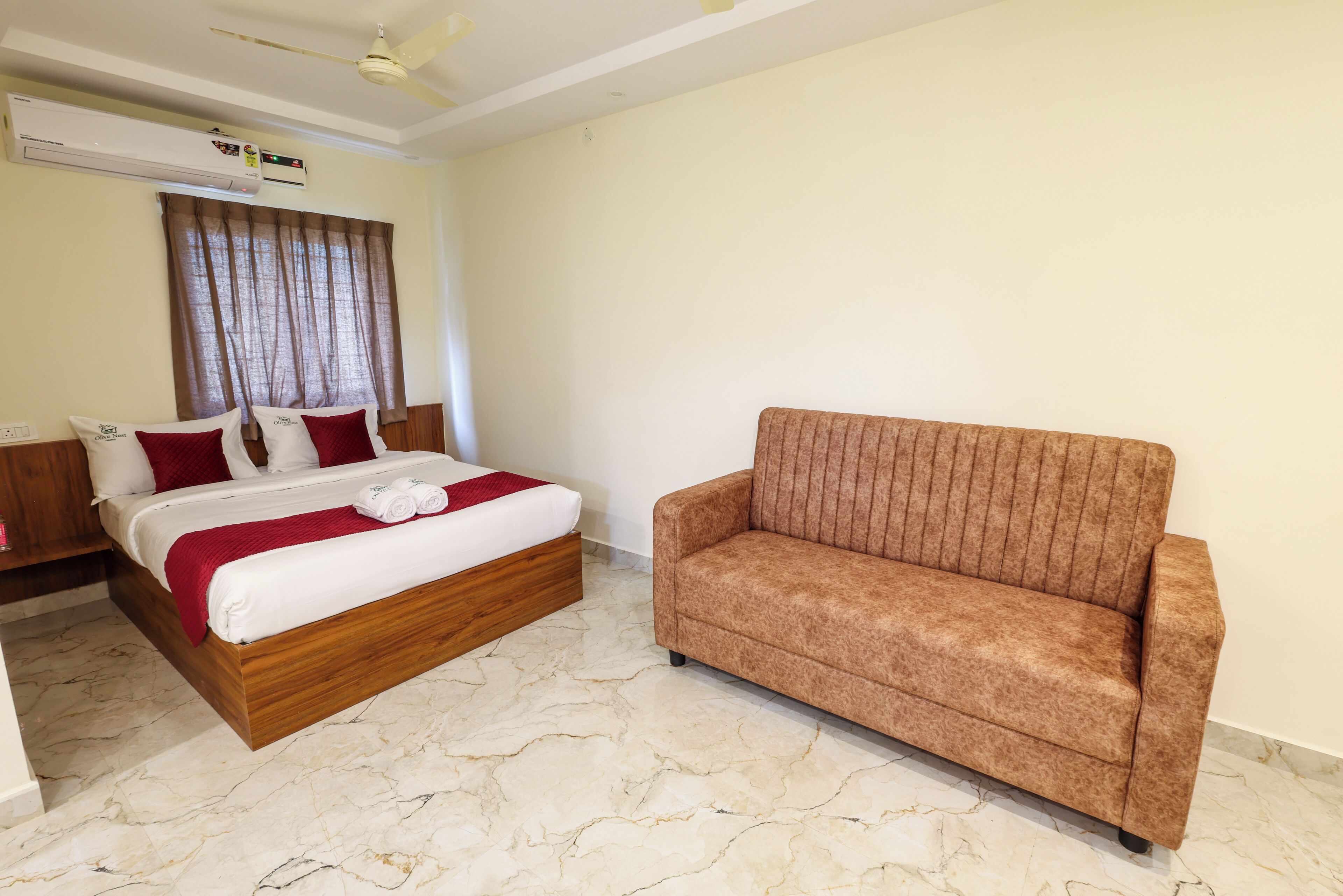 A hotel room with a bed, a brown sofa, a ceiling fan, and an air conditioning unit.