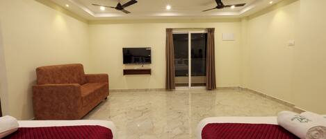A hotel room with two beds, a sofa, a TV, ceiling fans, and a sliding glass door.