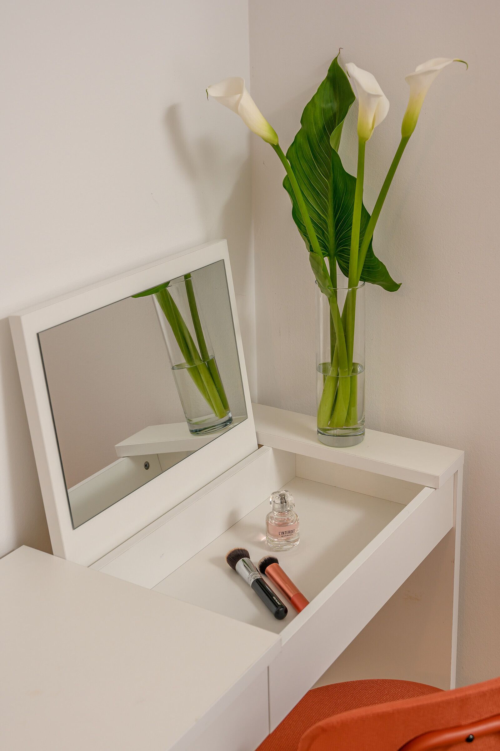 A white vanity with a mirror, a vase with white flowers, and a small bottle of perfume.