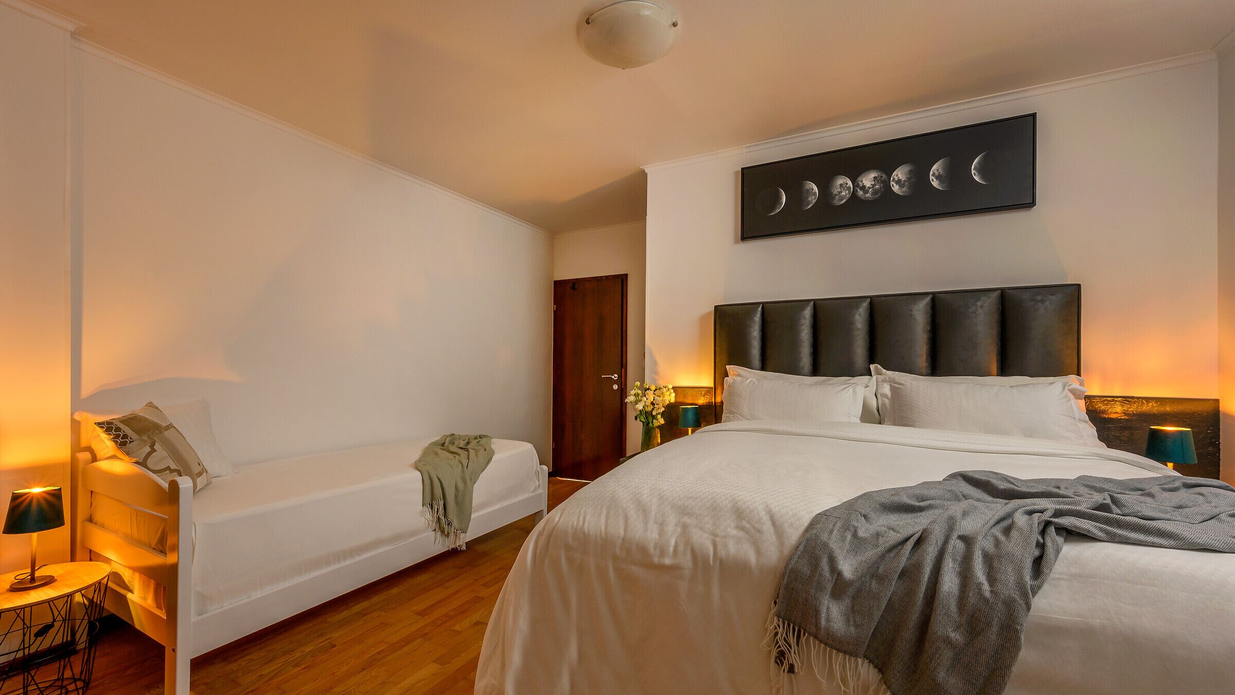 A hotel room with a large bed, a smaller bed, a nightstand with a lamp, a wall-mounted artwork depicting the moon phases, and a wooden floor.