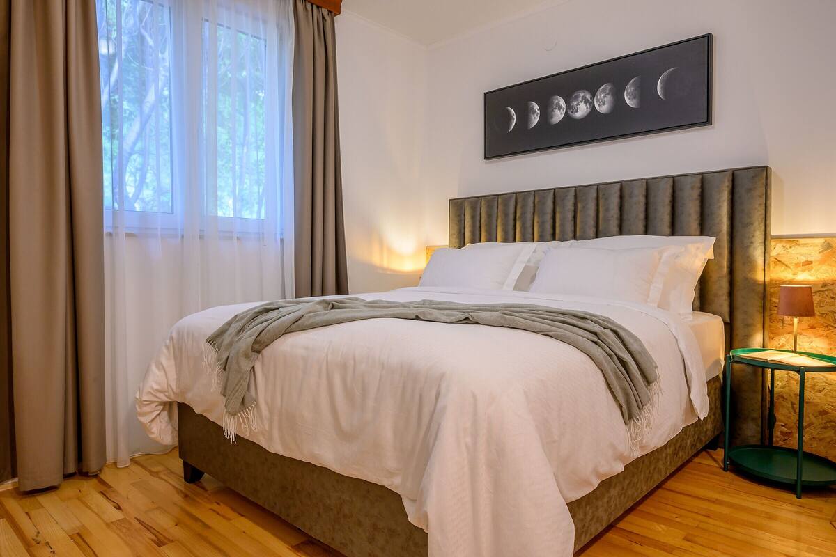 Una cama bien hecha con un plaid gris, una obra de arte con las fases de la luna enmarcada en la pared y una lámpara sobre una mesita de noche.