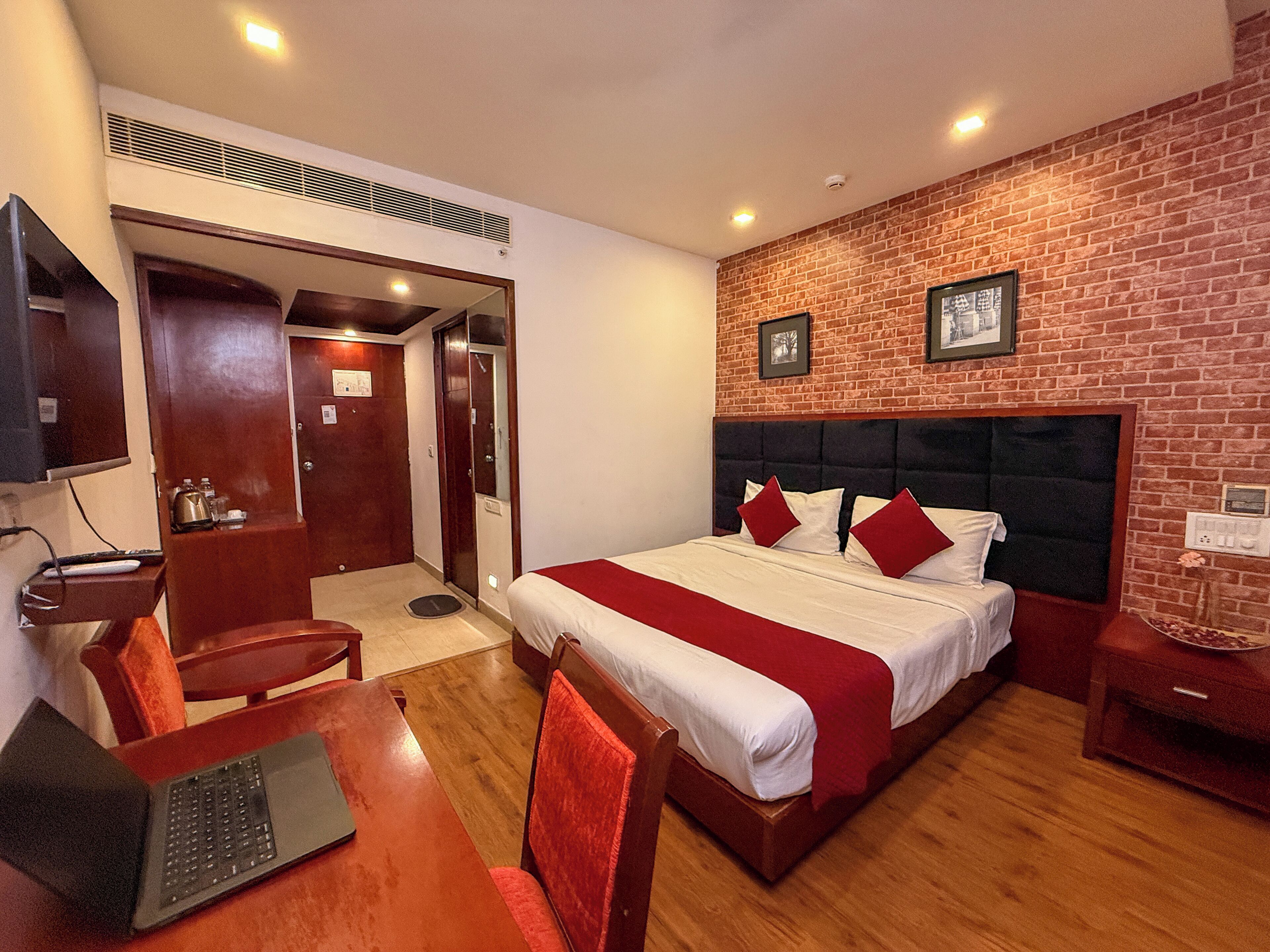 A hotel room with a bed, a desk with a laptop, a chair, a TV, and a brick wall.