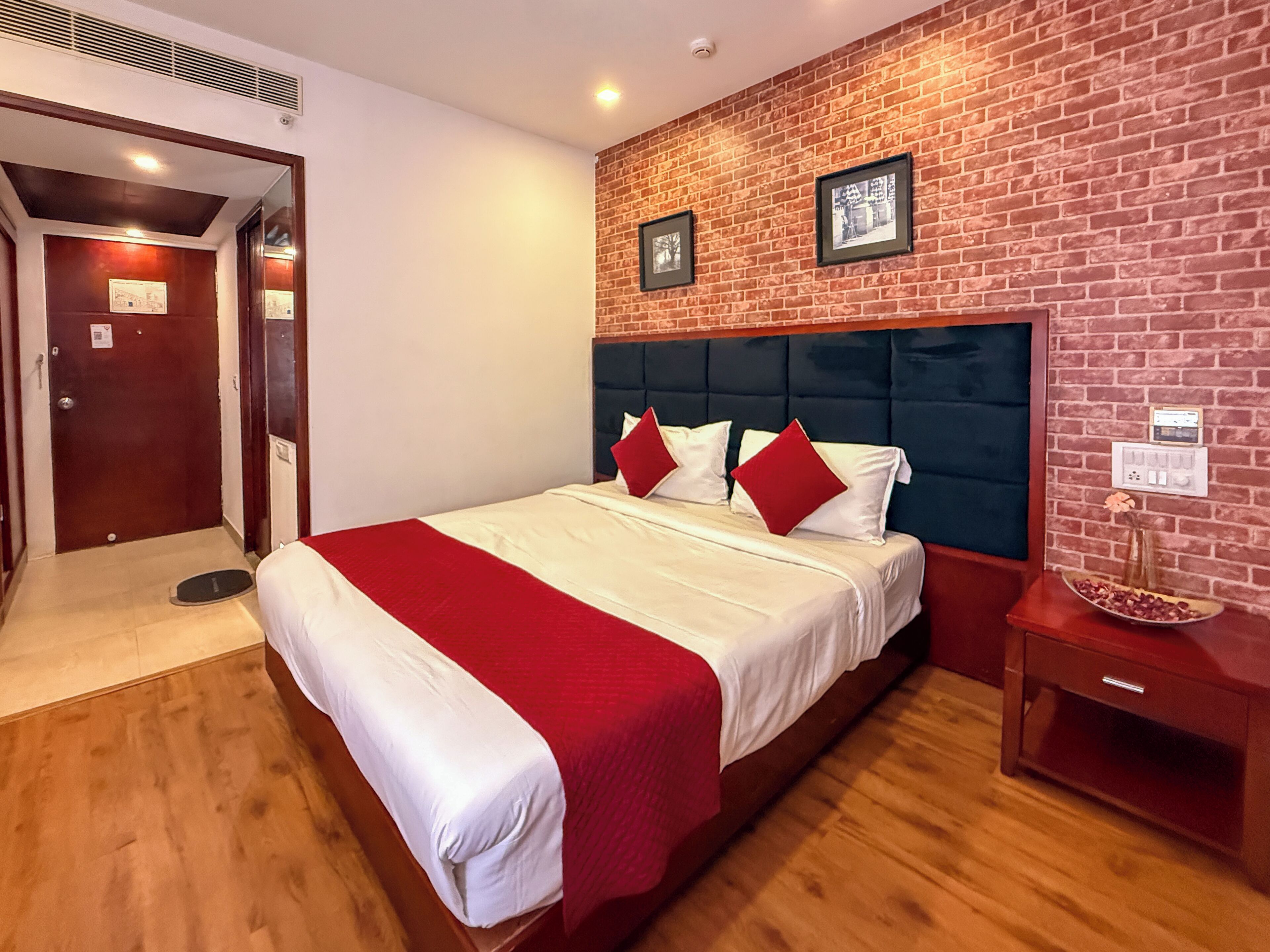 Une chambre d’hôtel avec un mur décoré d’un motif en briques, un grand lit avec une literie rouge et blanche, un plancher en bois et une table de chevet sur laquelle se trouve un vase.