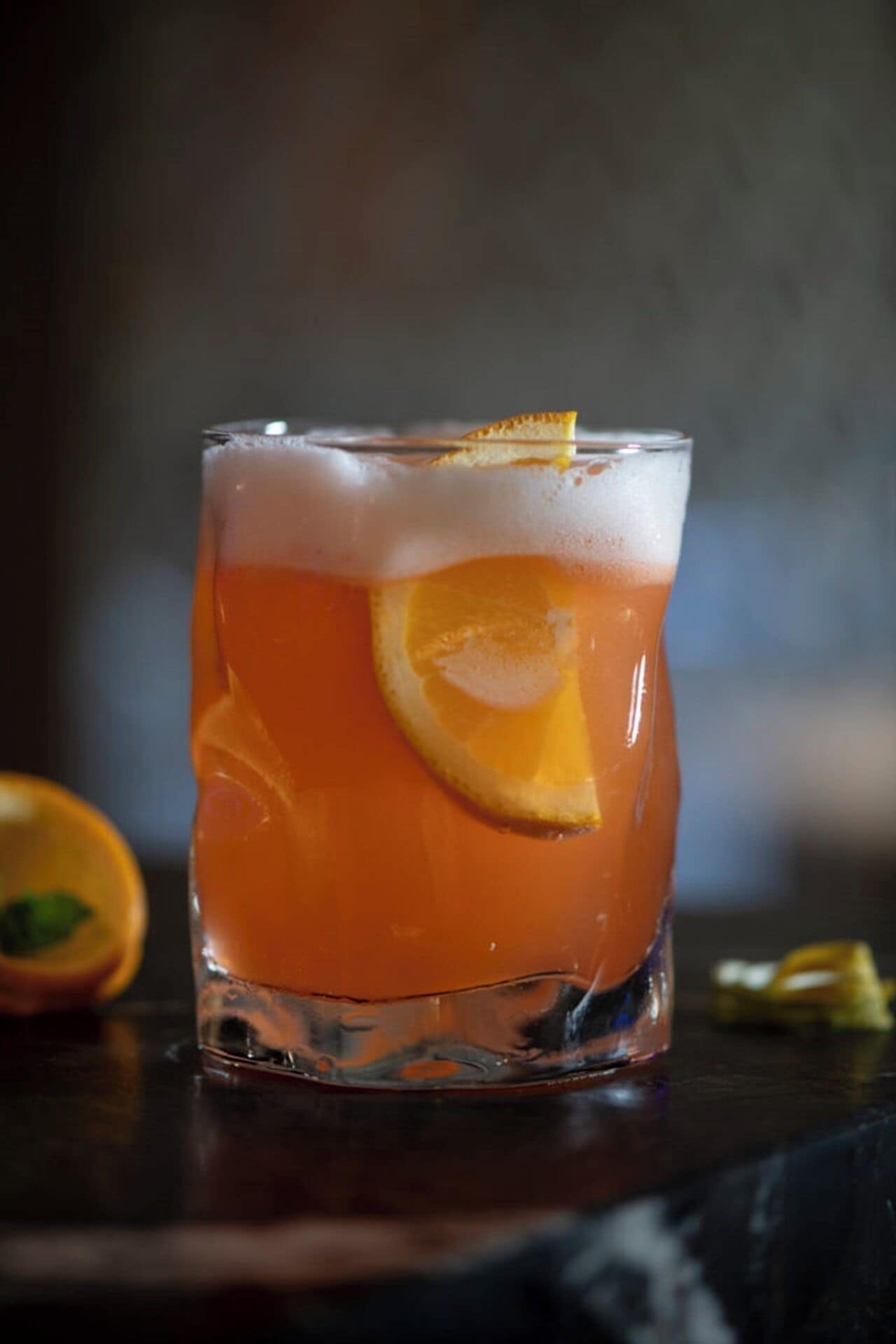 Een glas met een oranje drank, een oranjeschijfje en een citroenrasp, geplaatst op een donker oppervlak met een onscherpe achtergrond.