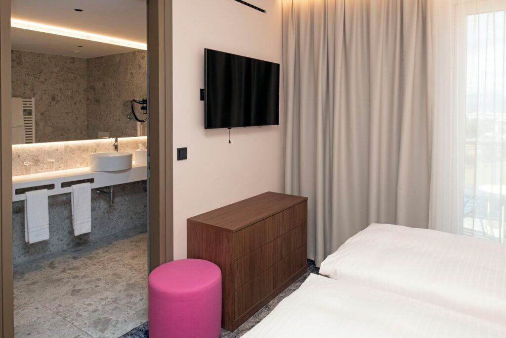 A modern hotel room with a large bed, a wooden dresser, a flat-screen TV mounted on the wall, a bathroom with a marble countertop, and a large window with curtains.