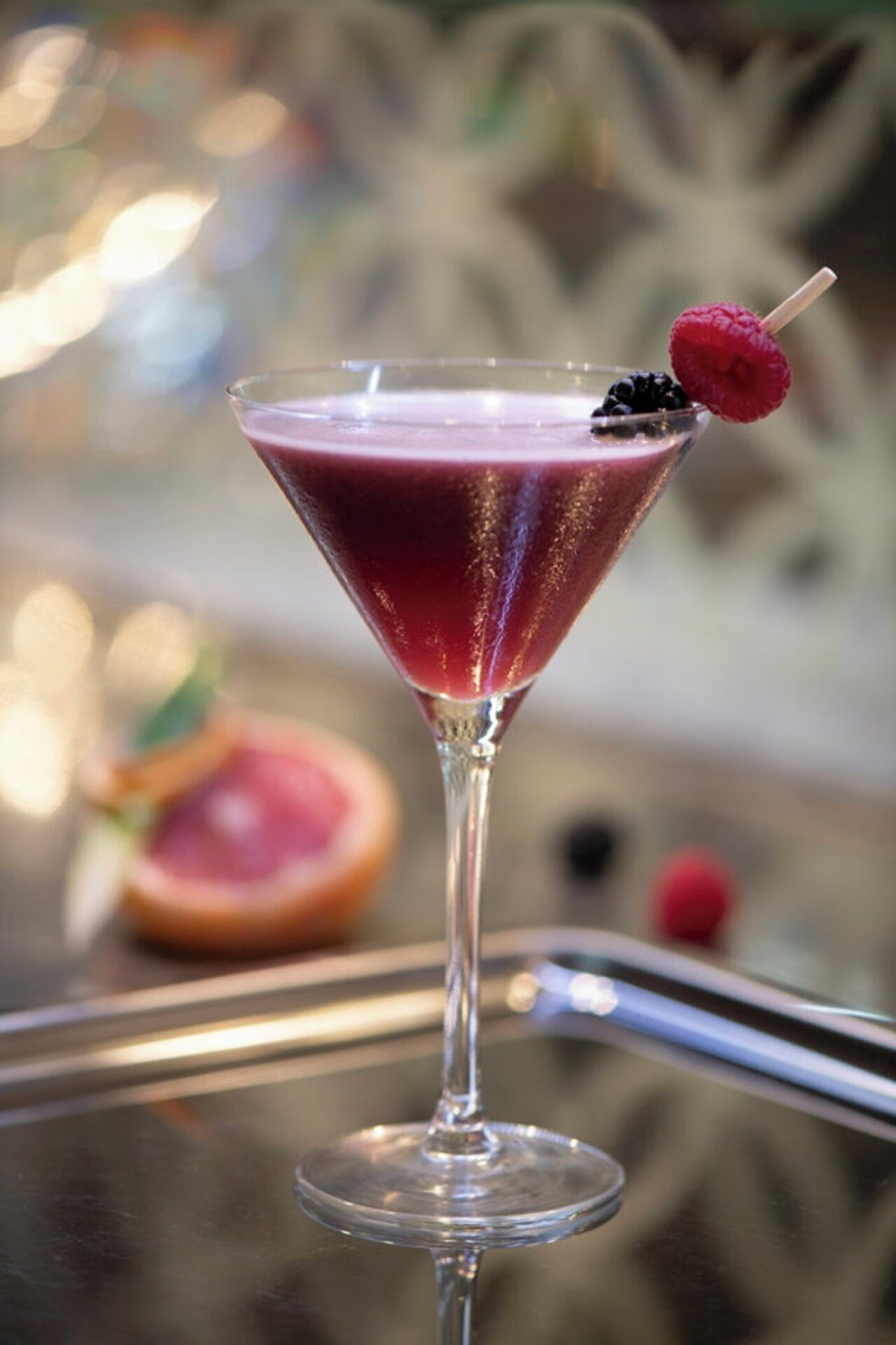 Una copa de martini con una bebida rosa, adornada con una frambuesa en un palillo, y un fruto cortado de fondo.