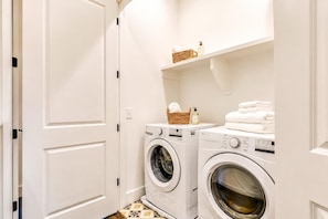 A laundry room with two front-loading washing machines, a shelf with folded towels, and a basket with a bottle.