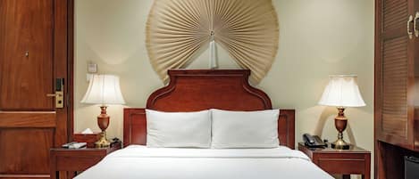 A hotel room with a large bed, two bedside tables with lamps, a wooden wardrobe, and a wall-mounted fan.