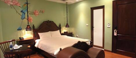 Habitación de hotel con una cama grande, un cabecero de madera, un escritorio con silla, una mesita pequeña y una lámpara de pared.
