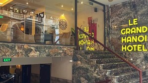En hotellobby med en marmortrappa, en kristallkrona och en skylt med texten ”LE GRAND HANOH HOTEL”.
