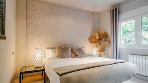A bedroom with a large bed, a bedside table with a lamp, and a view of greenery through a window.