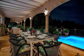 An outdoor dining area with a pool, surrounded by columns and potted plants.