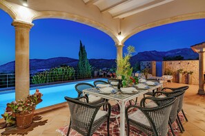 An outdoor dining area with a pool, surrounded by stone walls and potted plants, overlooking a mountainous landscape.