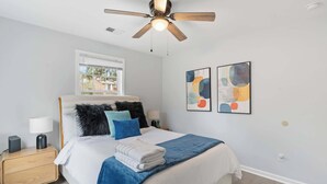 Un dormitorio con cama, mesita de noche, ventilador de techo, ventana y dos cuadros abstractos en la pared.