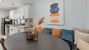 Una cocina moderna con armarios blancos, una mesa de comedor con un jarrón de flores secas y un cuadro abstracto colorido en la pared.