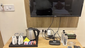 A hotel room with a wooden desk, a television, a coffee maker, a kettle, a power strip, a remote control, a water bottle, a glass, and a small refrigerator. - Hotel Kings Residency (Ahmedabad)