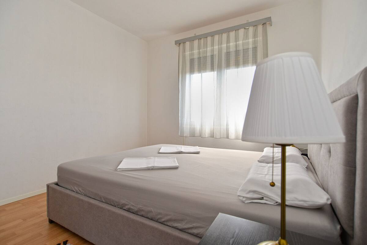 A minimalist bedroom with a single bed, a bedside lamp, and a window with sheer curtains.