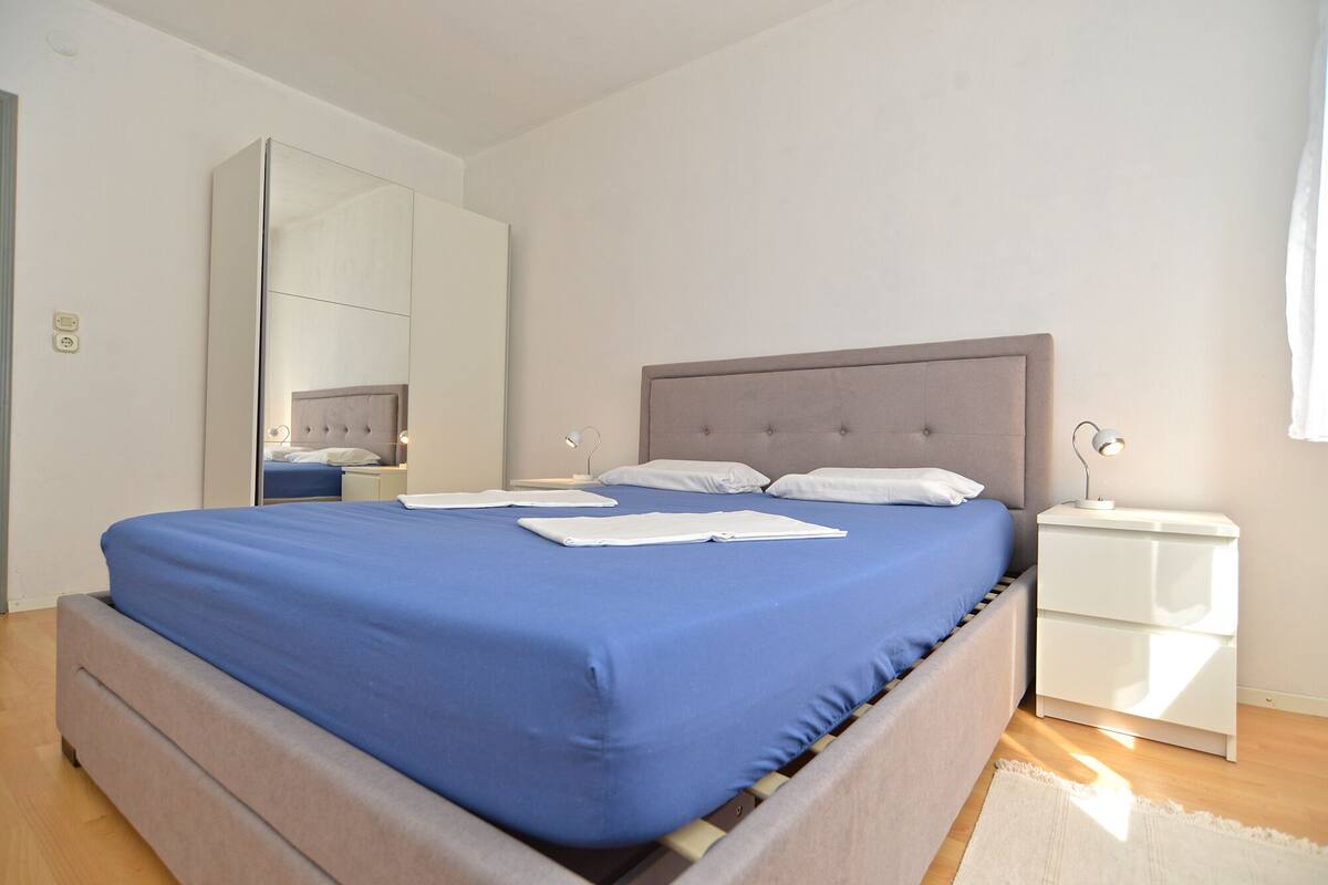 A modern bedroom with a large bed, a bedside table, a nightstand, a mirror, and a white wardrobe.