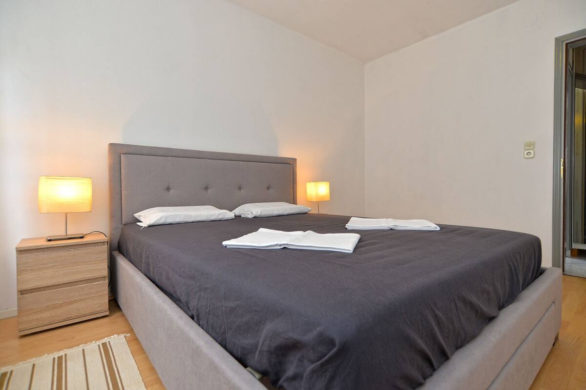 A neatly made bed with a grey headboard and two bedside lamps, a wooden nightstand, and a folded white towel on the bed.
