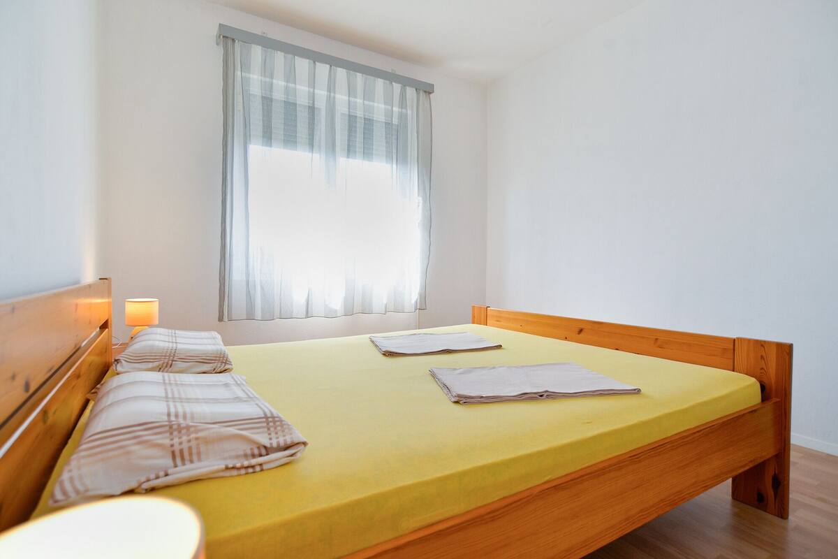 A single-bed room with a wooden headboard, a yellow bedspread, and a window with sheer curtains.