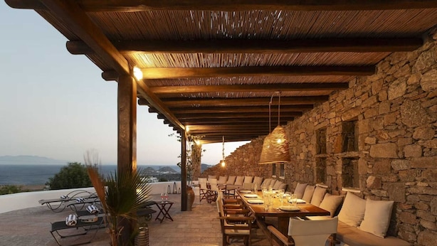 An outdoor dining area with stone walls, wooden furniture, and a view of the sea.