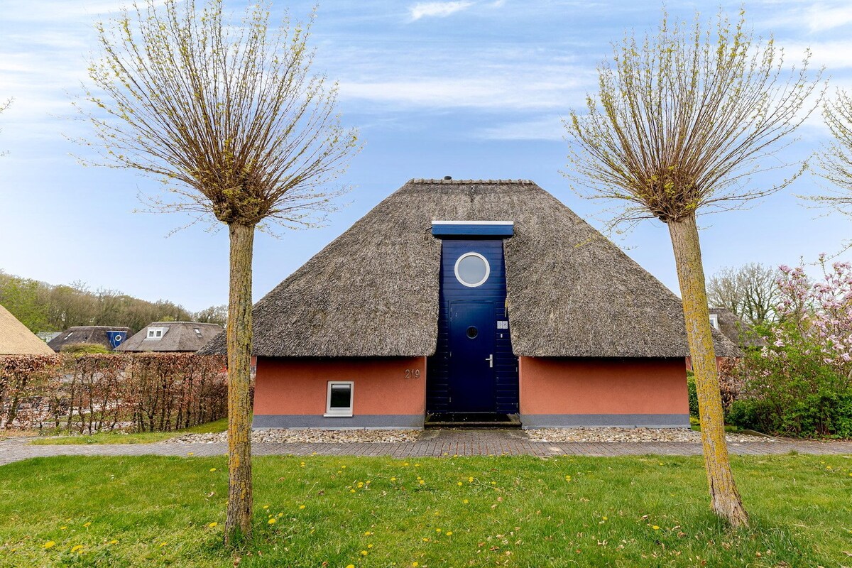 Une maison traditionnelle à toit de chaume, avec une porte bleu foncé, entourée d’une pelouse bien entretenue et de deux arbres.
