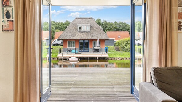 A view from a room with a wooden deck leading to a house with a thatched roof, a small dock, and a pond. - Summio Waterpark De Bloemert (Midlaren)