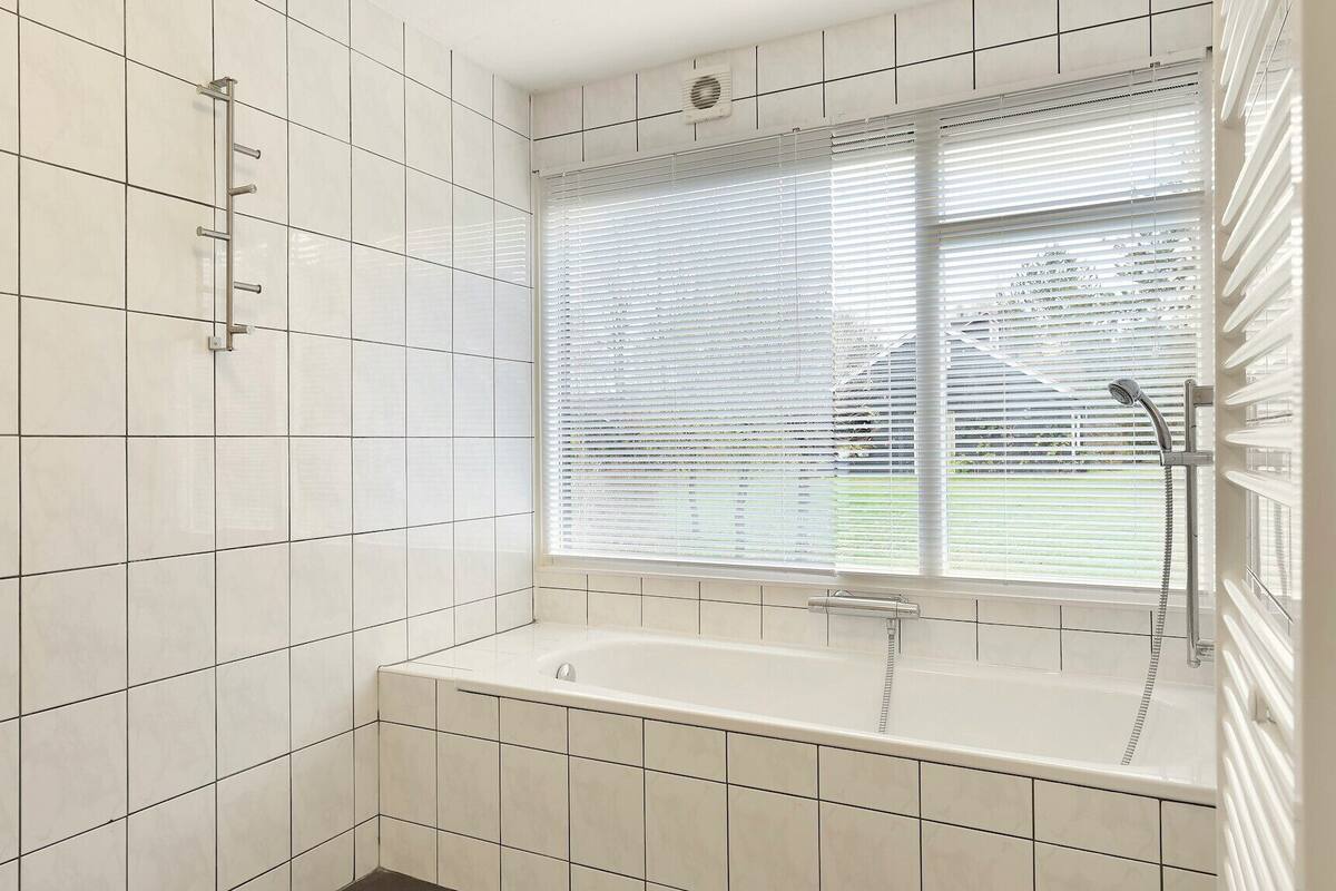Une salle de bain moderne avec des carreaux blancs, une baignoire, une fenêtre avec des stores et une pomme de douche.
