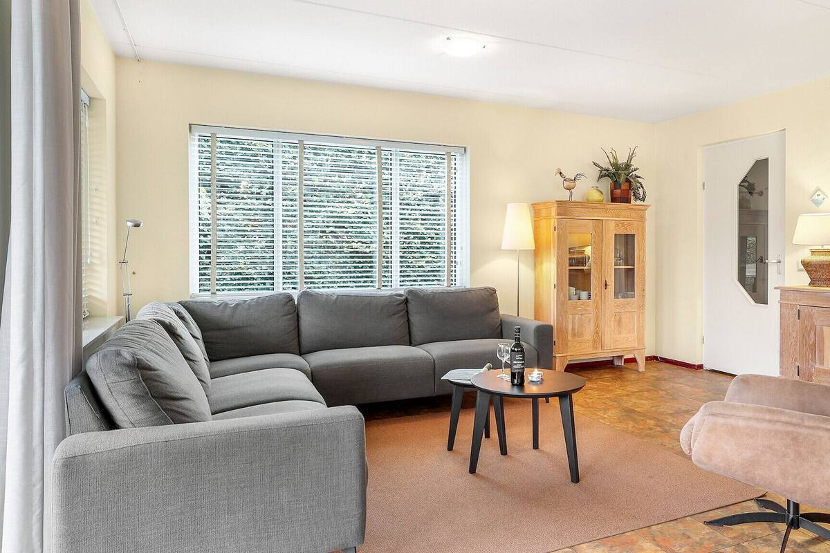 Un salon avec un canapé gris, une petite table ronde sur laquelle se trouvent une bouteille et un verre, un meuble en bois, un fauteuil beige et un téléviseur posé sur un meuble en bois.
