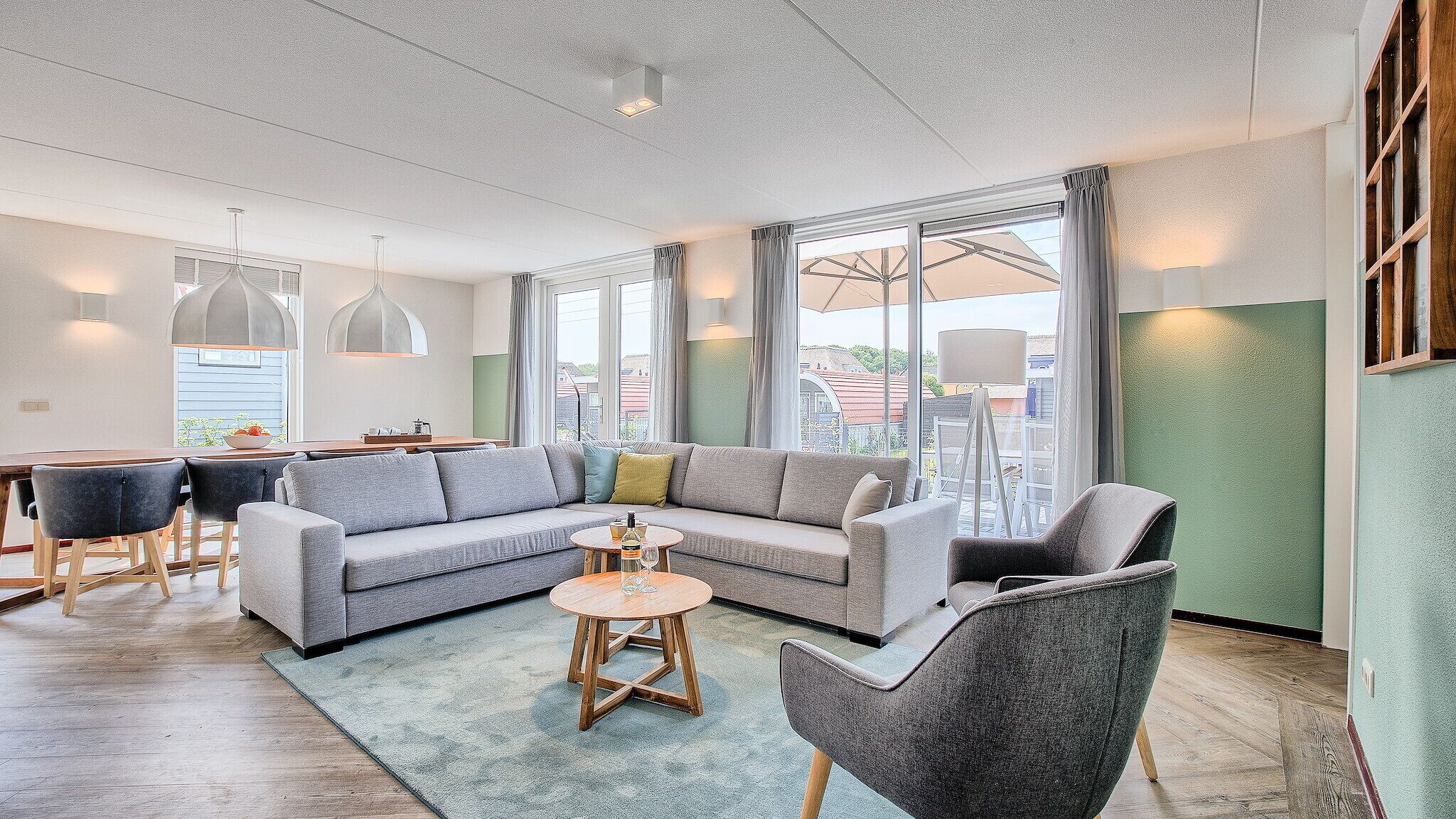 Un salon moderne comprenant un canapé, des fauteuils, une table basse et un coin repas avec une table et des chaises. De grandes fenêtres laissent entrer la lumière naturelle et offrent une vue sur l’extérieur.