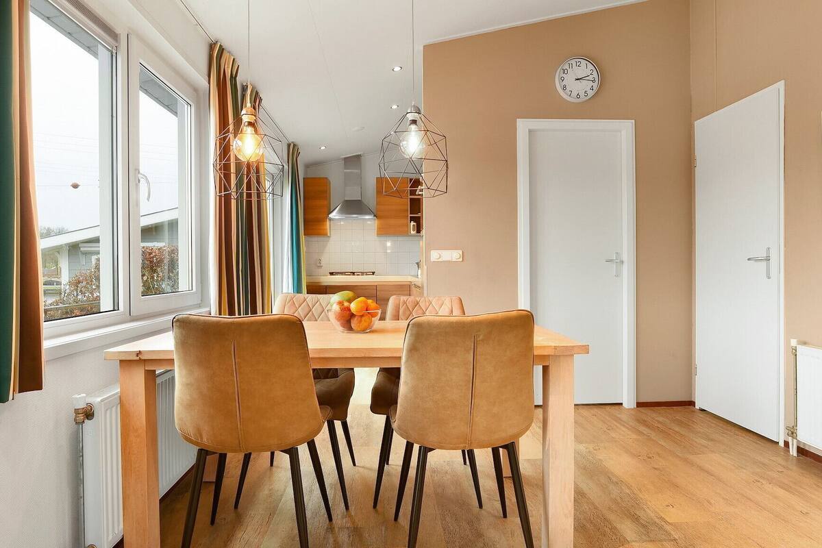 Un coin repas avec une table en bois et deux chaises, un panier de fruits sur la table, et une cuisine en arrière-plan.