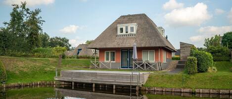 A thatched-roof house with a wooden dock over a calm water body, surrounded by greenery and other similar houses.