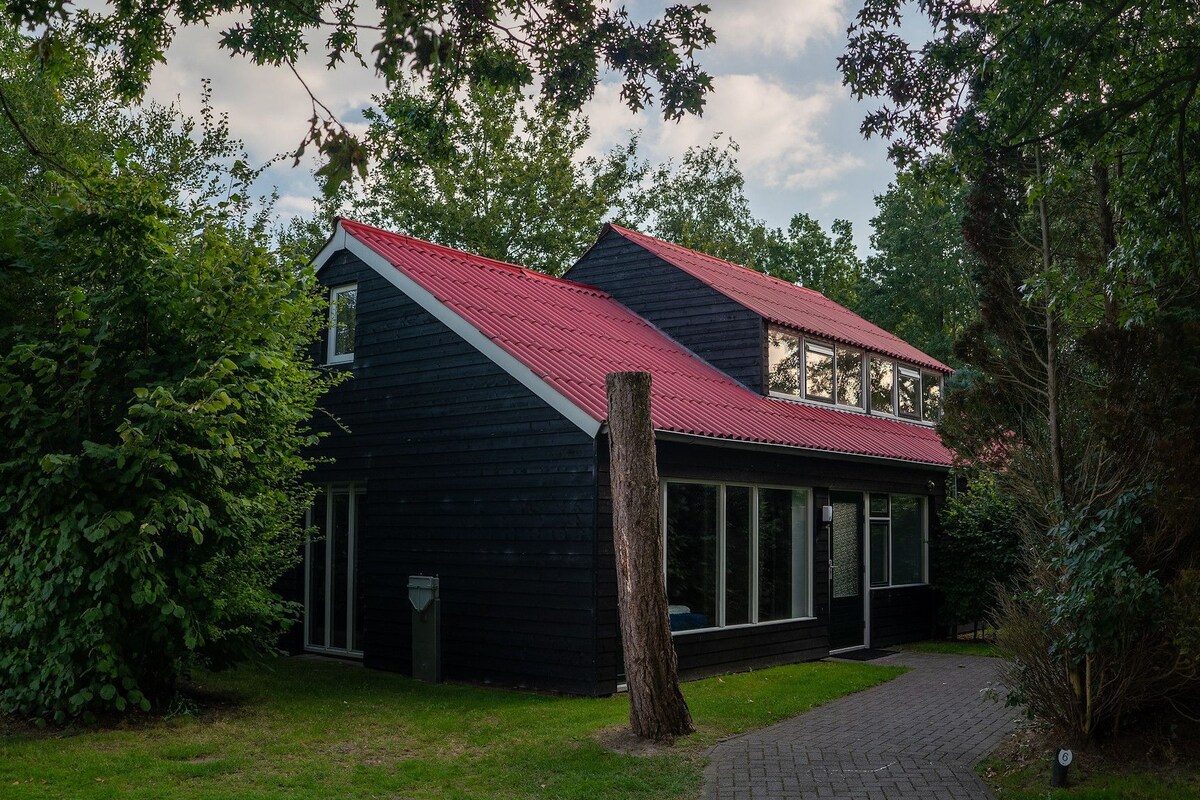 Une maison à deux étages, avec un toit rouge, entourée d’une végétation luxuriante.