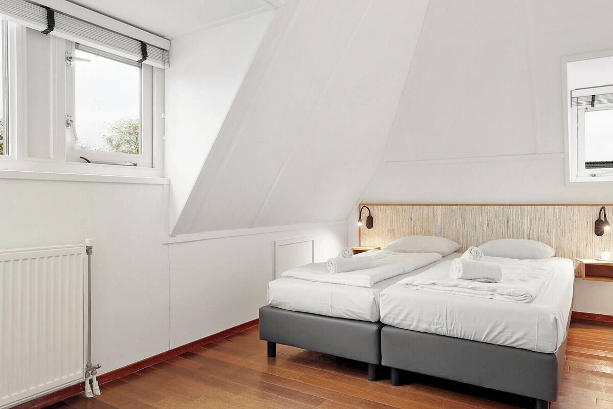 Une chambre moderne avec un plafond en pente, deux lits, une tête de lit en bois, des lampes de chevet et une vue sur l’extérieur.