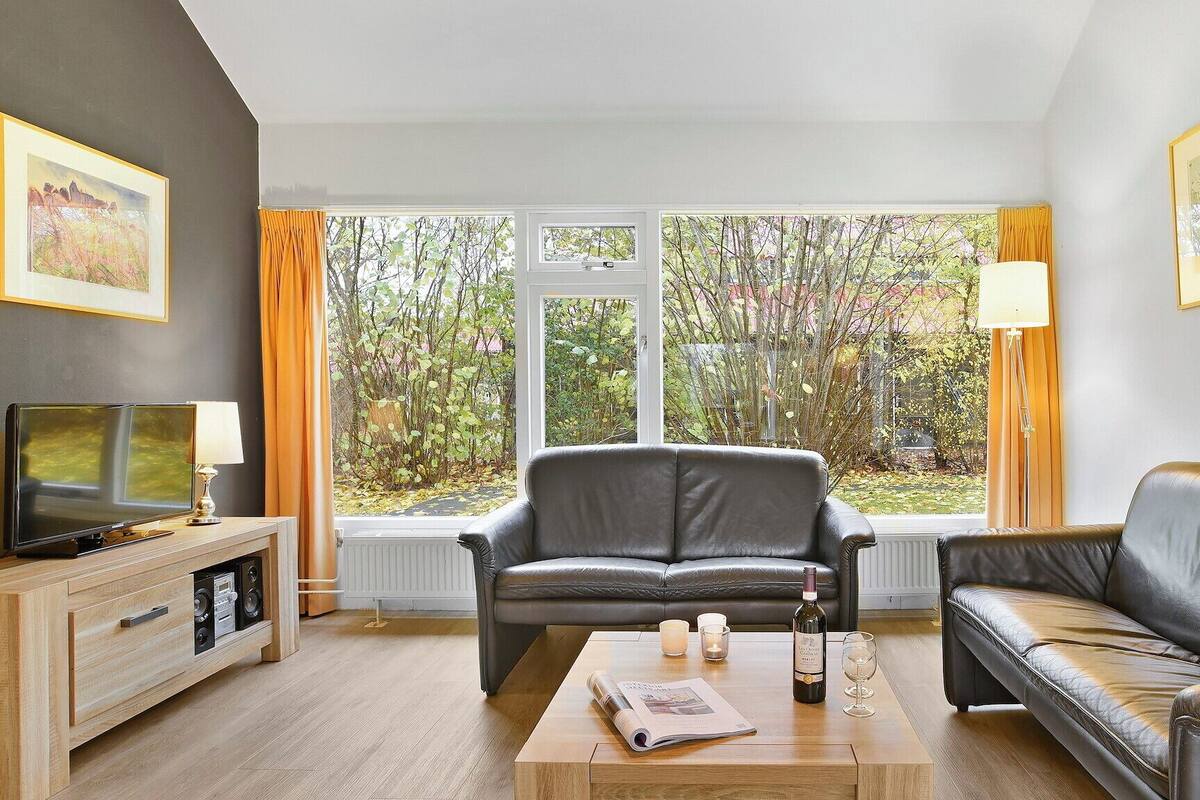 Un salon avec un canapé en cuir, un téléviseur, une table basse sur laquelle repose un journal et une fenêtre avec des rideaux.