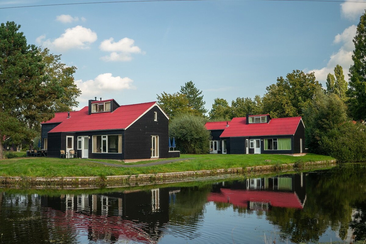 Deux maisons noires aux toits rouges, situées au bord d’un lac paisible, et entourées d’arbres.