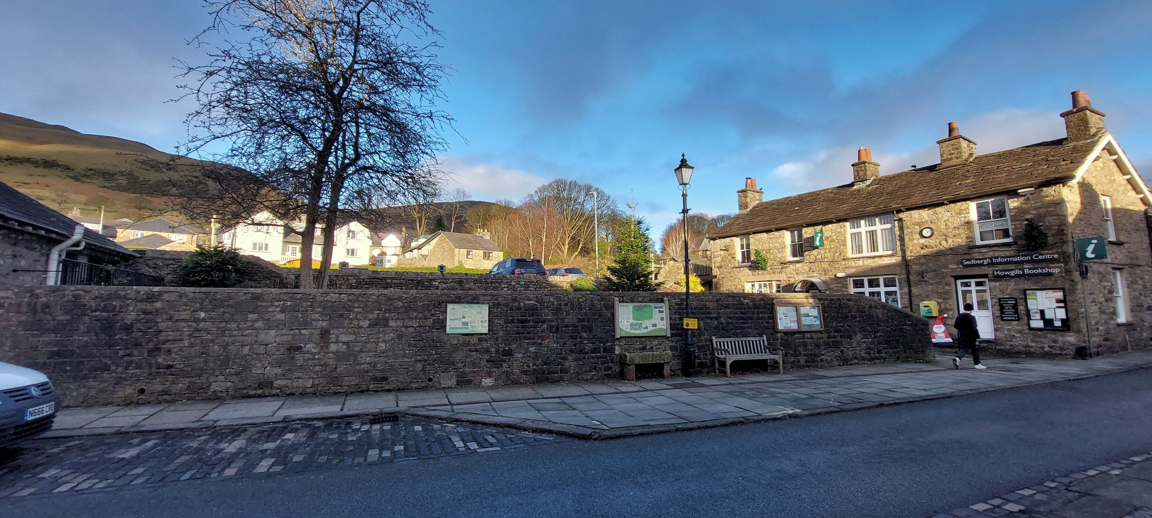 Un muro de piedra con bancos y carteles informativos, un edificio de piedra con un letrero que dice 'Howgills Bookshop', y un coche estacionado.