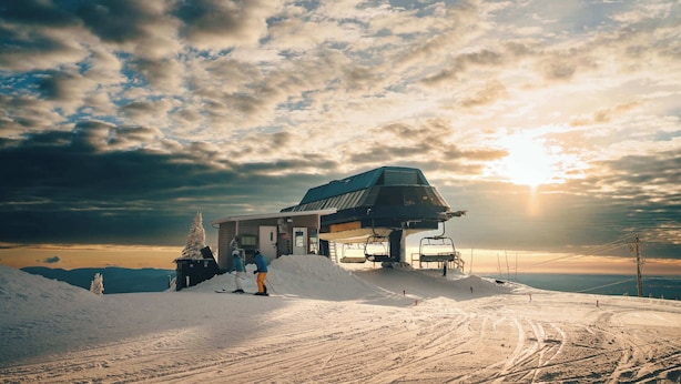 Estación de telesilla con un ascensor de sillones, suelo cubierto de nieve y un edificio con tejado inclinado.