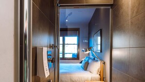 Una habitación de hotel moderna con cabina de ducha de vidrio, una cama con almohadas, una mesita de noche con lámpara y una ventana con luz natural.