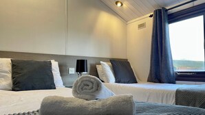 Habitación de hotel con dos camas, una ventana con cortinas, una lámpara fijada a la pared y una toalla enrollada sobre la cama.