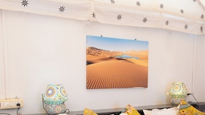 A hotel room with a bed, pillows, a nightstand, a lamp, and a wall art of a desert landscape. - Desert Fortune Resort (Jaisalmer)