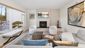 A modern living room with a fireplace, two white armchairs, a grey sofa, and a large abstract painting.