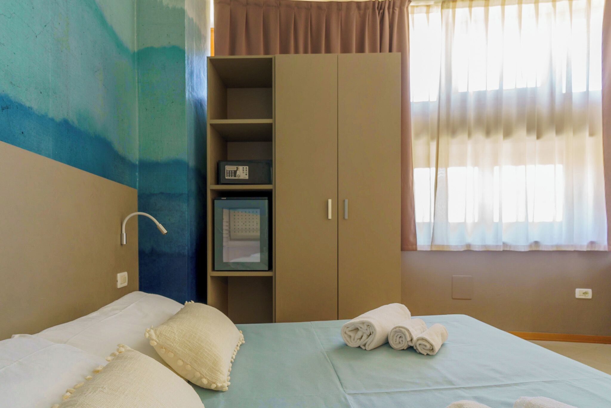 Een hotelkamer met een bed, een kledingkast, een plank met een radio en een raam met gordijnen.