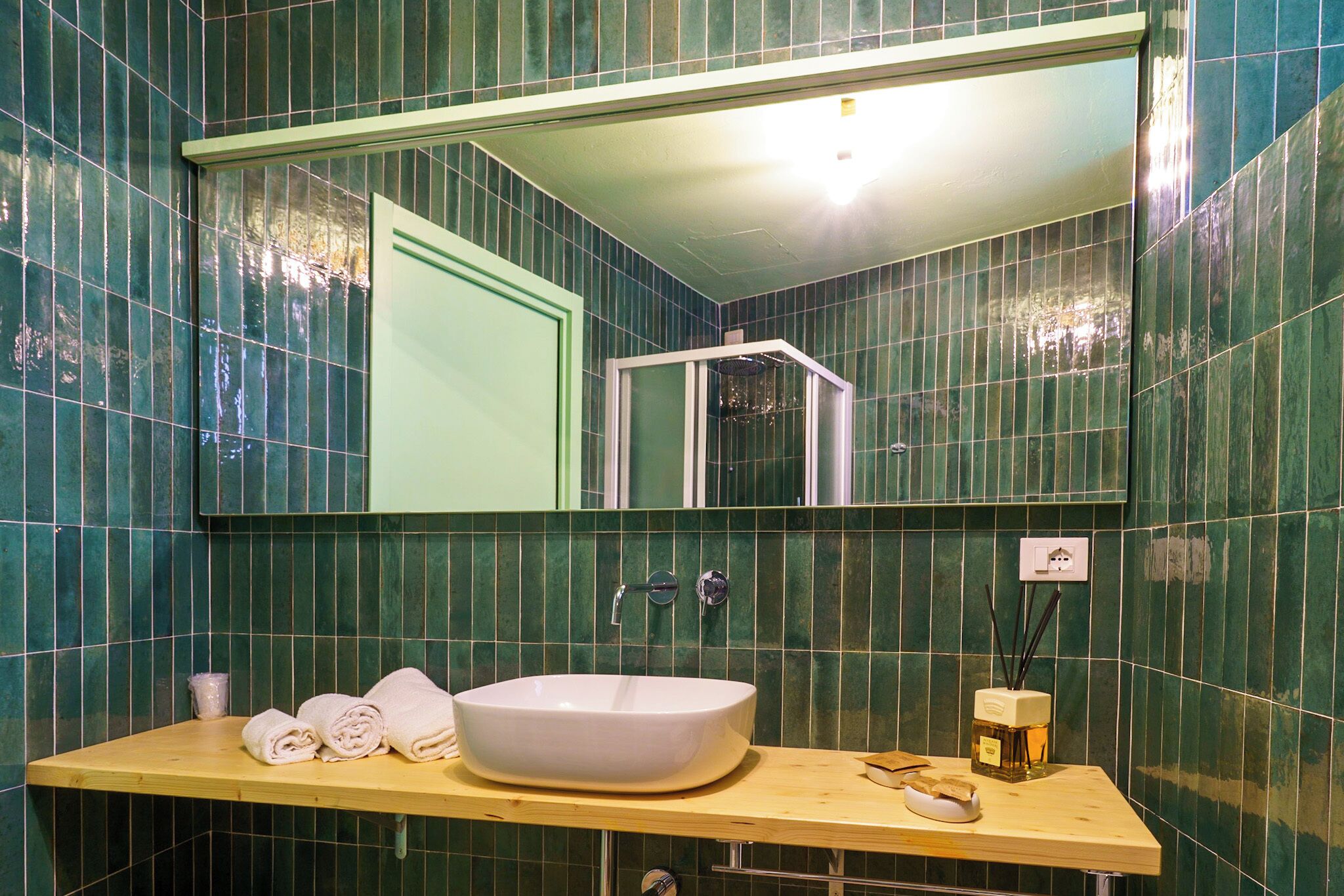 Un baño con un espejo grande, un tocador de madera, un lavamanos blanco y paredes cubiertas de azulejos verdes.